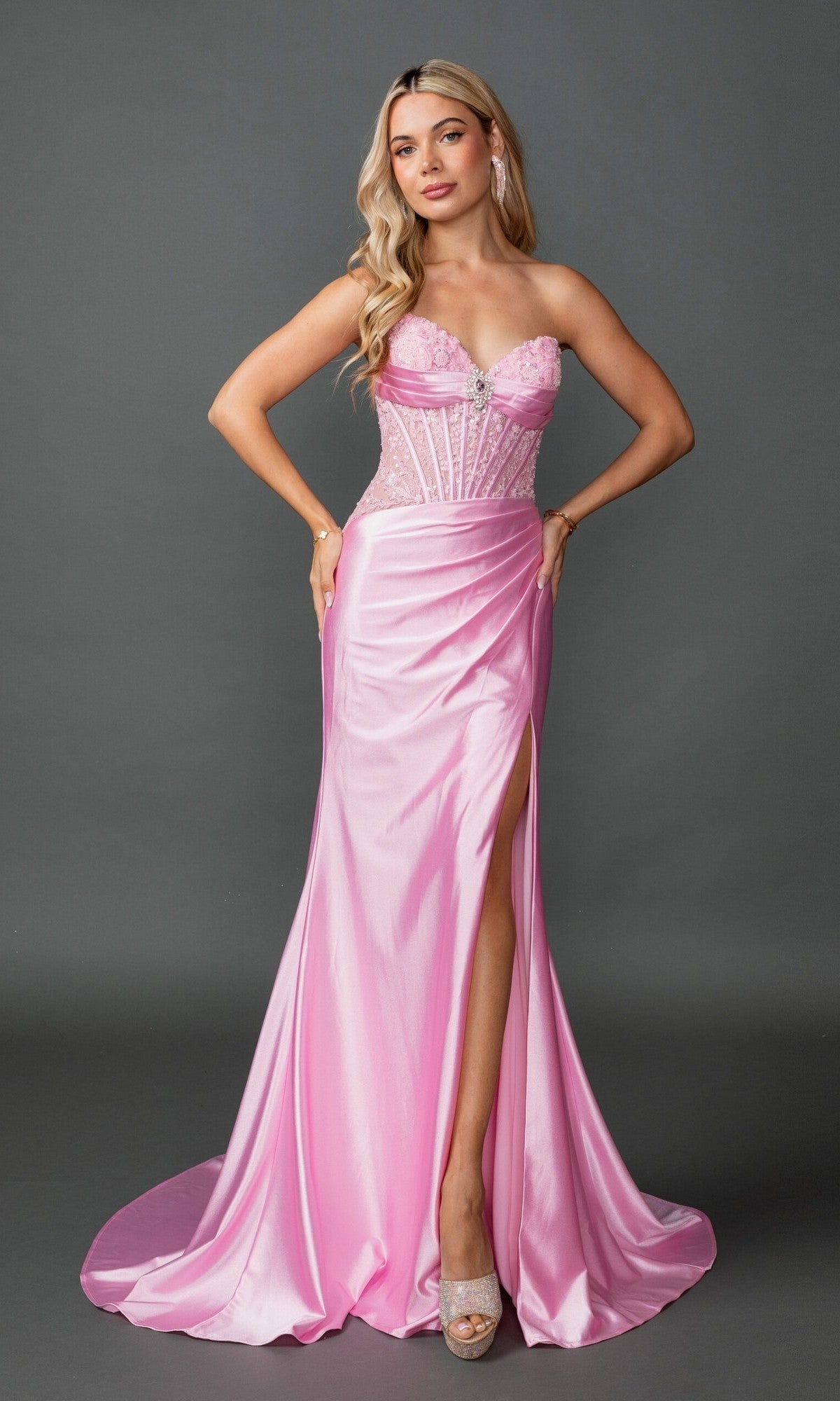A woman poses against a plain gray background wearing the Long Prom Dress: Dancing Queen 4587 in pink—a strapless satin gown featuring a sheer corset bodice with floral details, a fitted sheath skirt with a thigh-high slit, and paired with peep-toe heels.