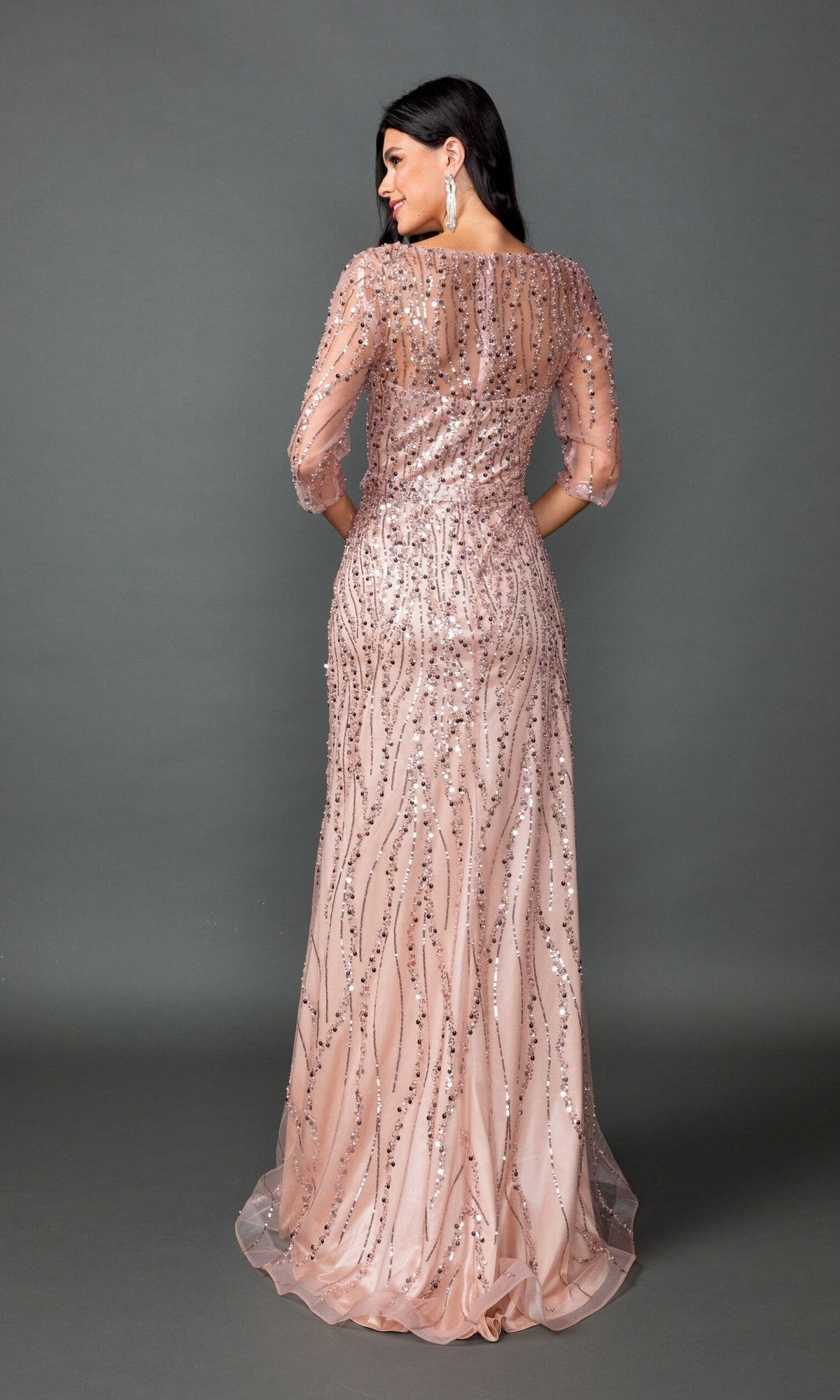 A woman stands with her back to the camera against a plain gray background, modeling the Dancing Queen 4588 long prom dress—a floor-length pink gown embellished with sequins and featuring sheer sleeves.