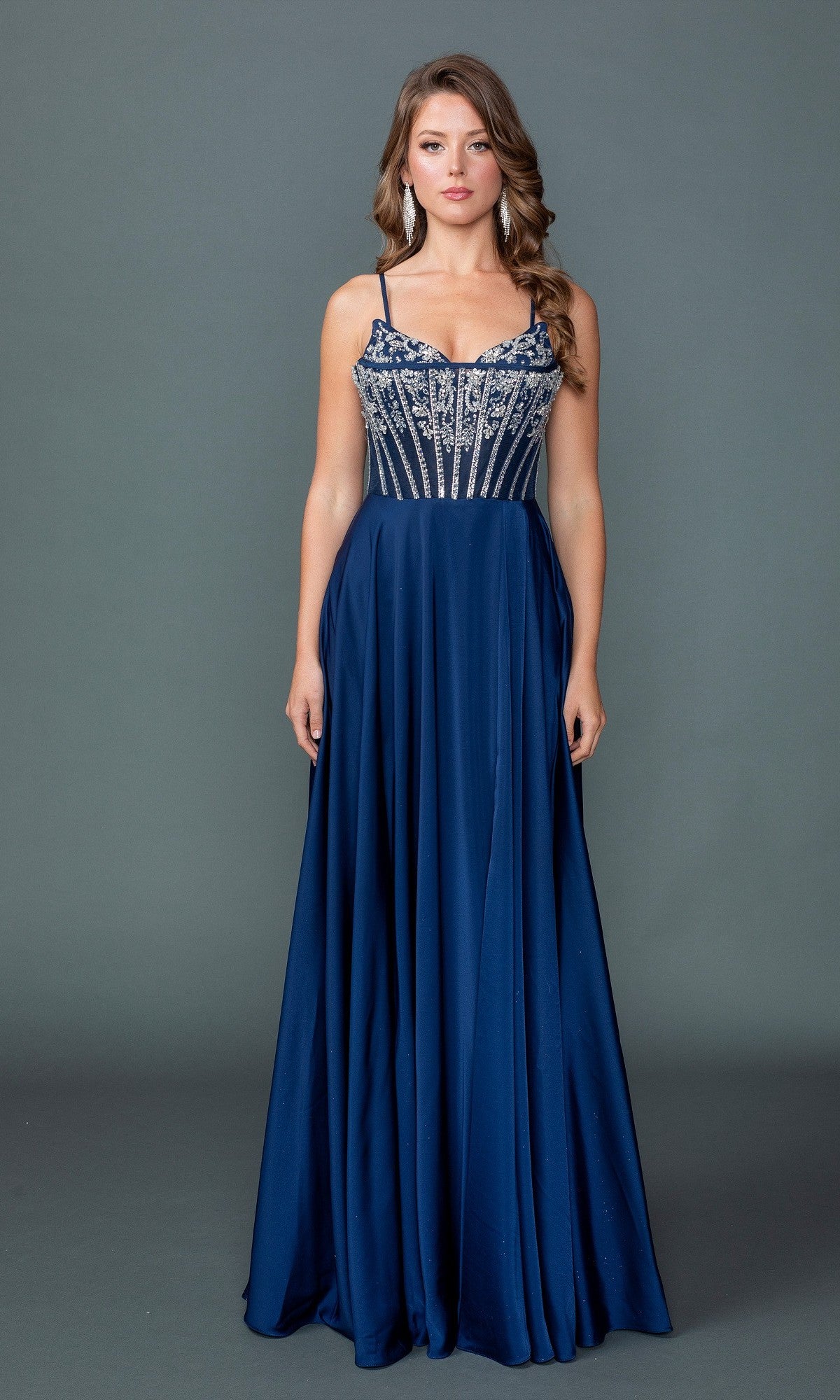 A woman poses against a gray background in the Long Prom Dress: Dancing Queen 4589, featuring a navy floor-length A-line silhouette with a beaded corset-style bodice, thin straps, and an elegant leg slit.