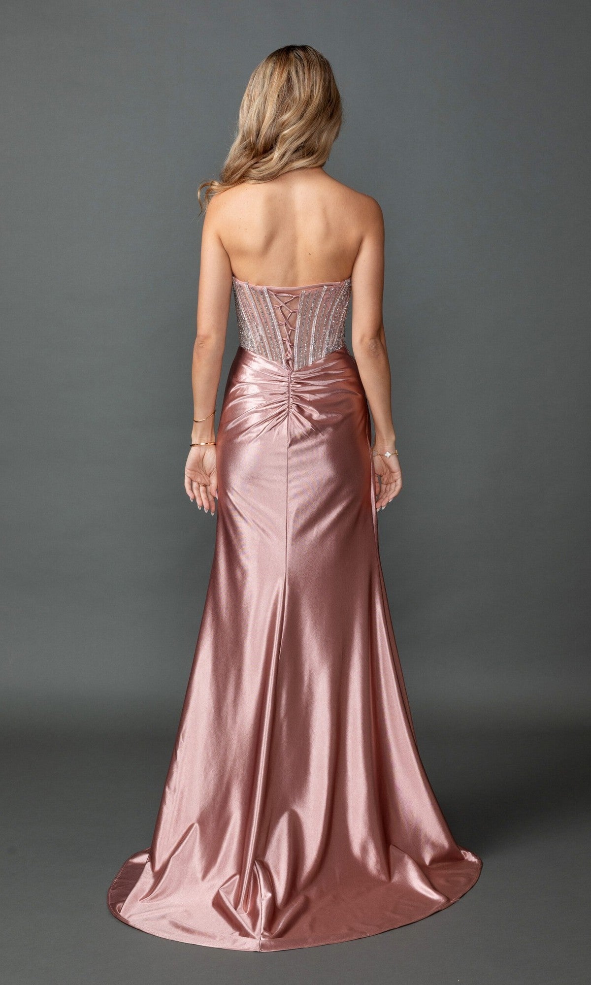 A woman stands with her back to the camera, wearing the Long Prom Dress: Dancing Queen 4593—a metallic pink, floor-length gown highlighted by a fitted, sheer corset bodice and ruched detailing on the back.