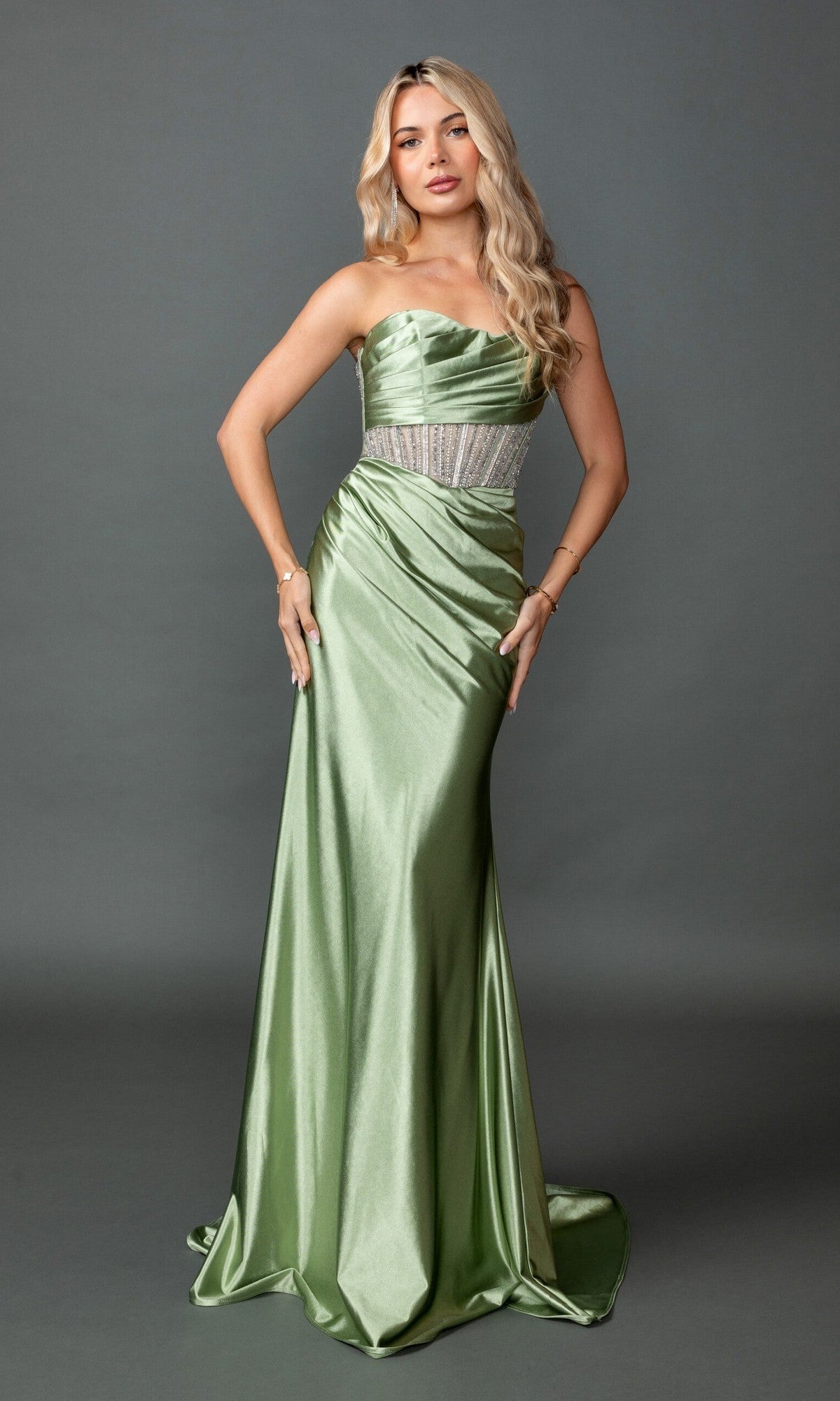 A woman stands against a gray background, wearing the Long Prom Dress: Dancing Queen 4593 in olive, which features a strapless design, jeweled belt, sheer corset bodice, long wavy blonde hair, and minimal jewelry.