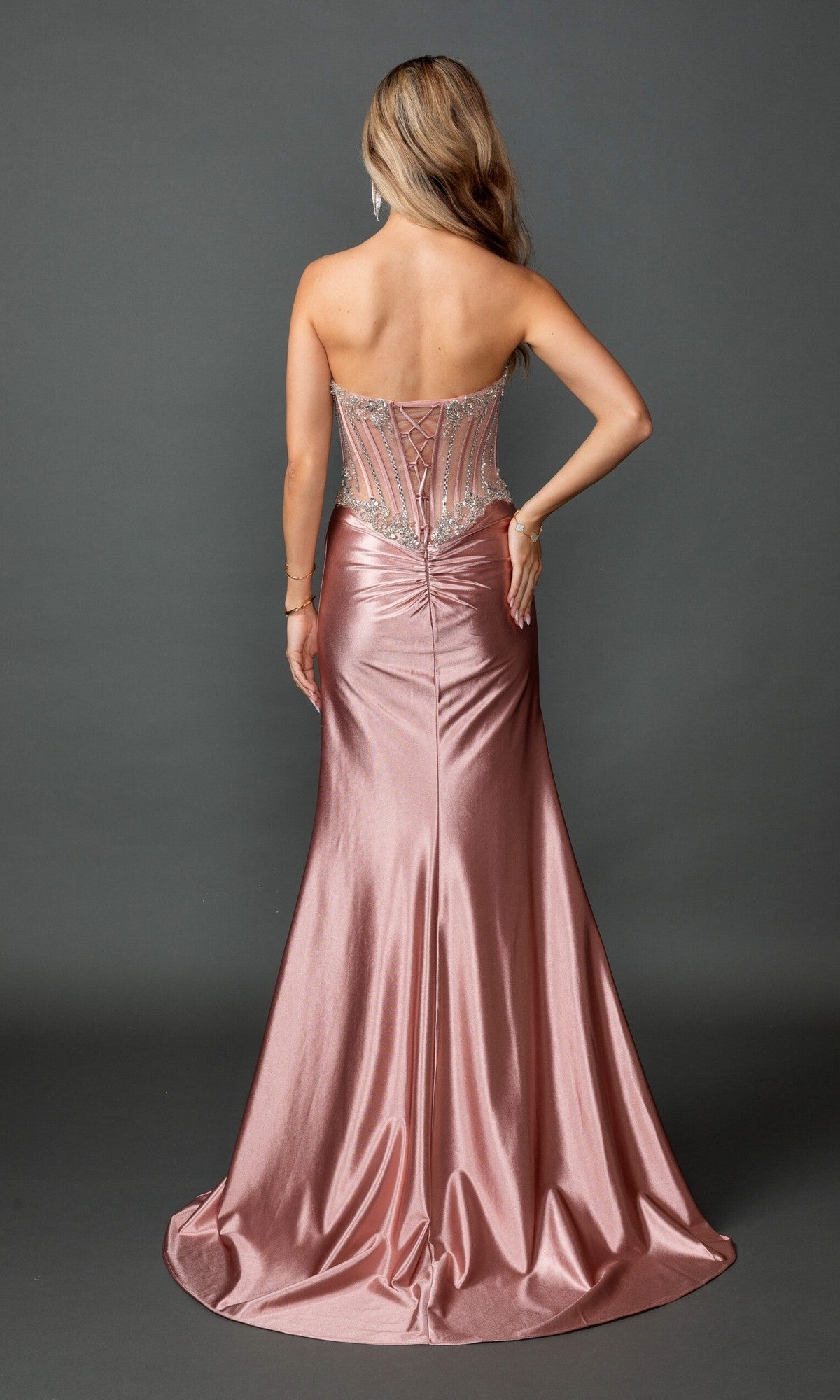 A woman stands facing away, wearing the Long Prom Dress: Dancing Queen 4594—a strapless metallic rose gold gown featuring a fitted, embellished corset bodice and a flared, floor-length skirt—set against a plain dark background.