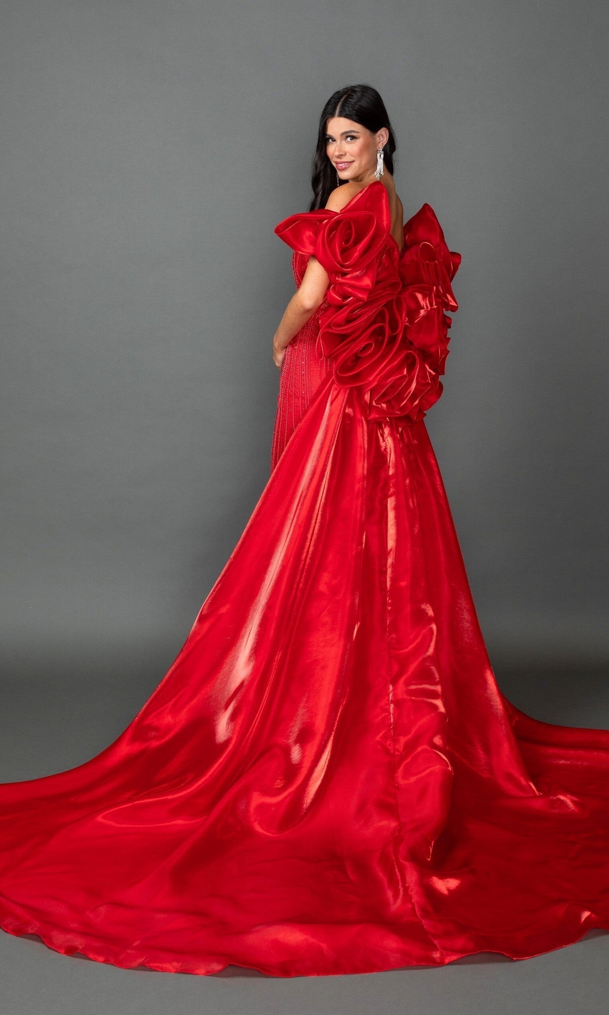 A woman wearing the Long Prom Dress: Dancing Queen 4597—a shiny red gown featuring an elaborate bow and floral detail on the back—stands against a plain gray background.