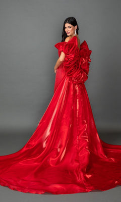 A woman wearing the Long Prom Dress: Dancing Queen 4597—a shiny red gown featuring an elaborate bow and floral detail on the back—stands against a plain gray background.