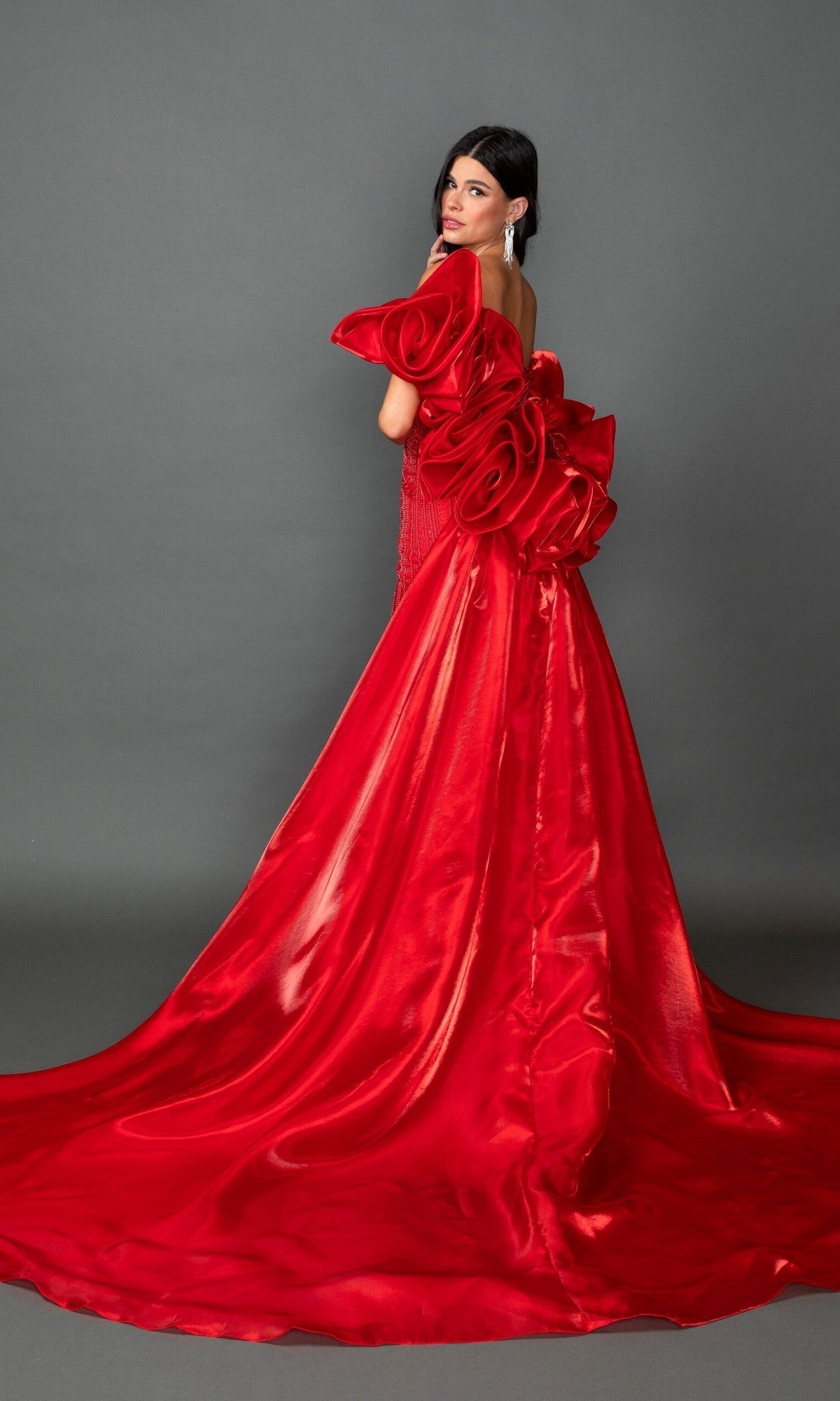 A woman wears the Long Prom Dress: Dancing Queen 4597, an elegant and shiny red strapless gown featuring dramatic floral ruffles on the back. She stands against a plain gray background, glancing over her shoulder with poise.