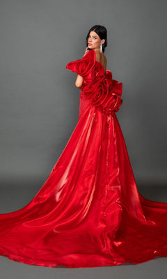 A woman wears the Long Prom Dress: Dancing Queen 4597, an elegant and shiny red strapless gown featuring dramatic floral ruffles on the back. She stands against a plain gray background, glancing over her shoulder with poise.