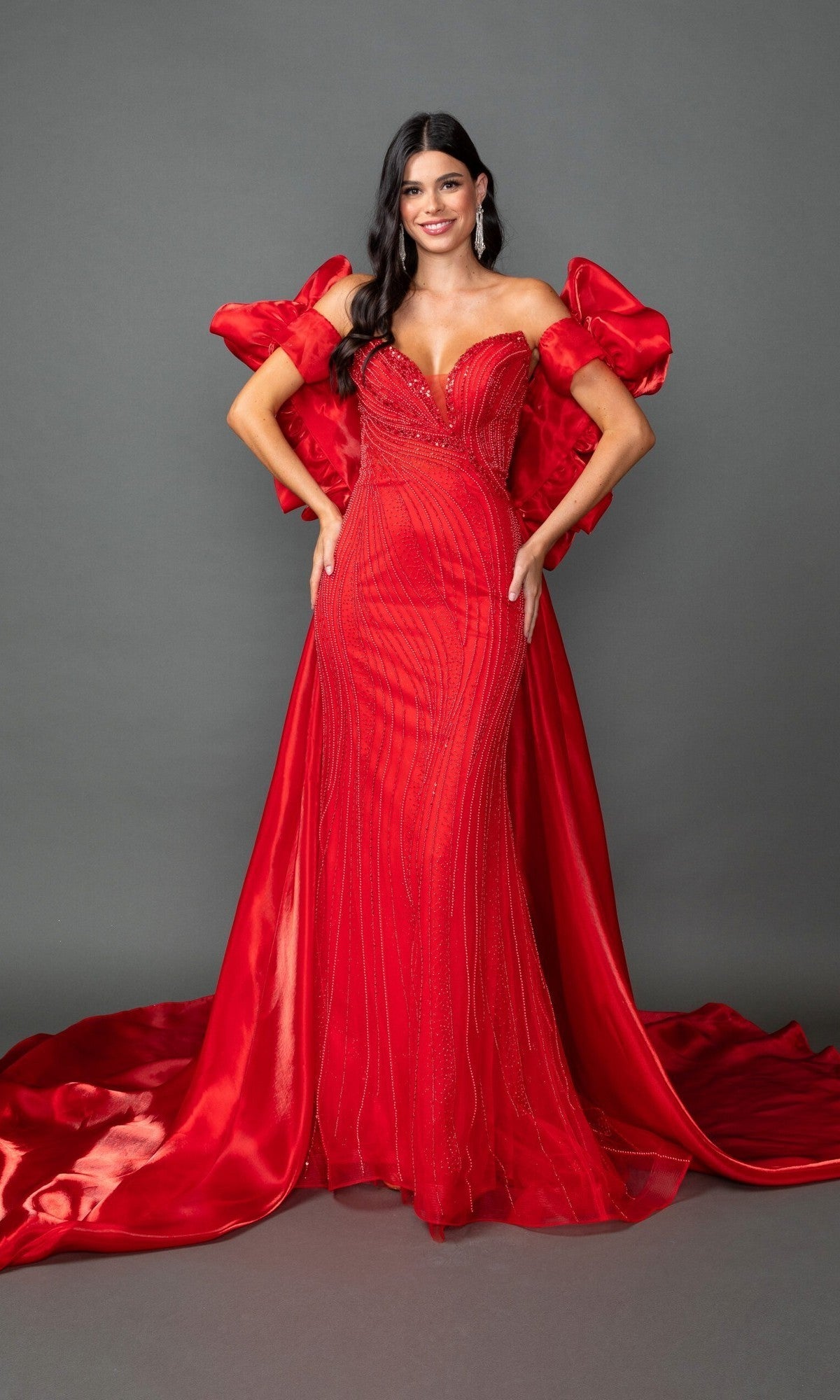 A woman stands in the Long Prom Dress: Dancing Queen 4597, a floor-length, fitted red gown featuring beaded details, dramatic puffed sleeves, and a sweeping train, set against a plain gray background.