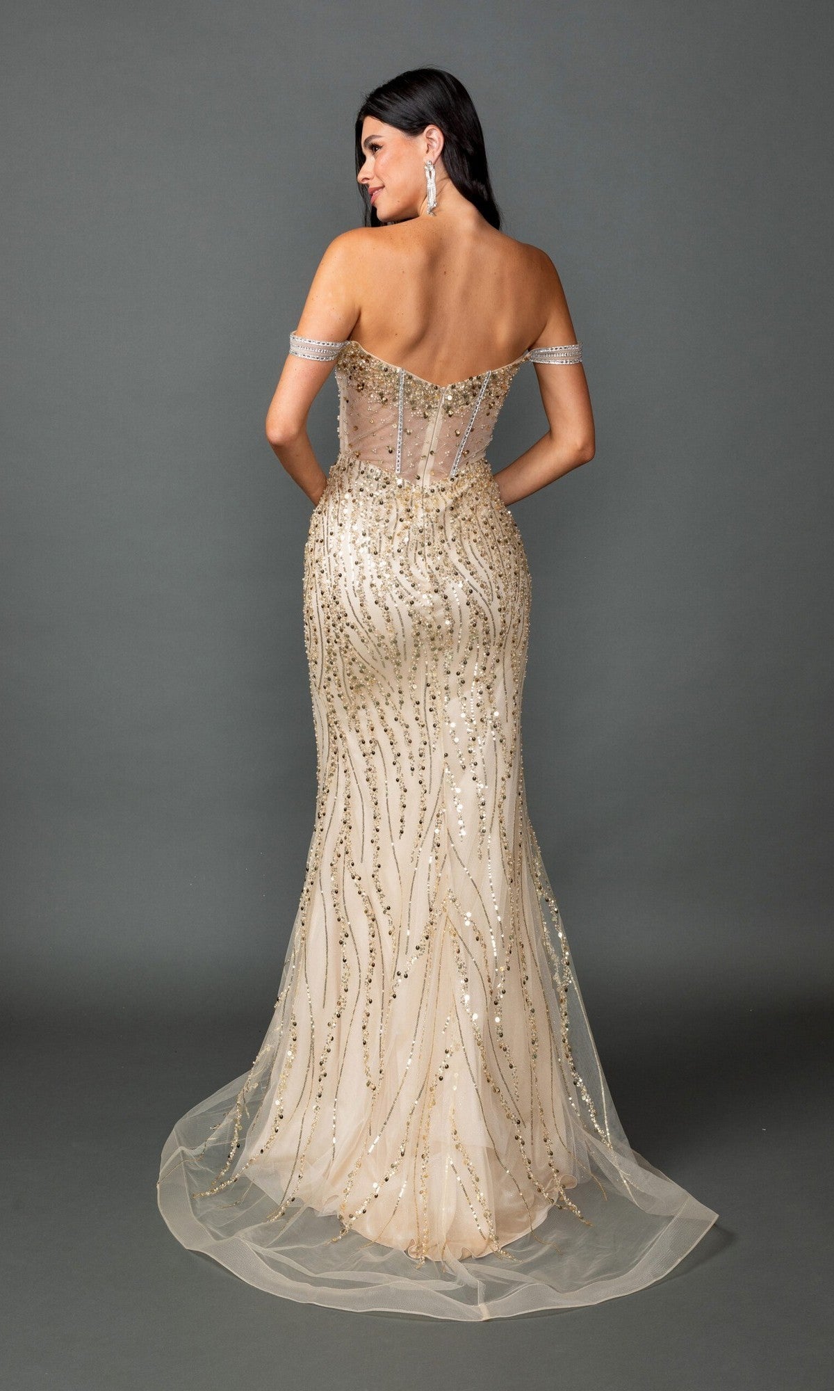 A woman wearing the Dancing Queen 4598 long prom dress stands facing away, showcasing its off-the-shoulder, floor-length silhouette with a sheer corset bodice, gold sequin embellishments, and decorative beading against a plain gray background.