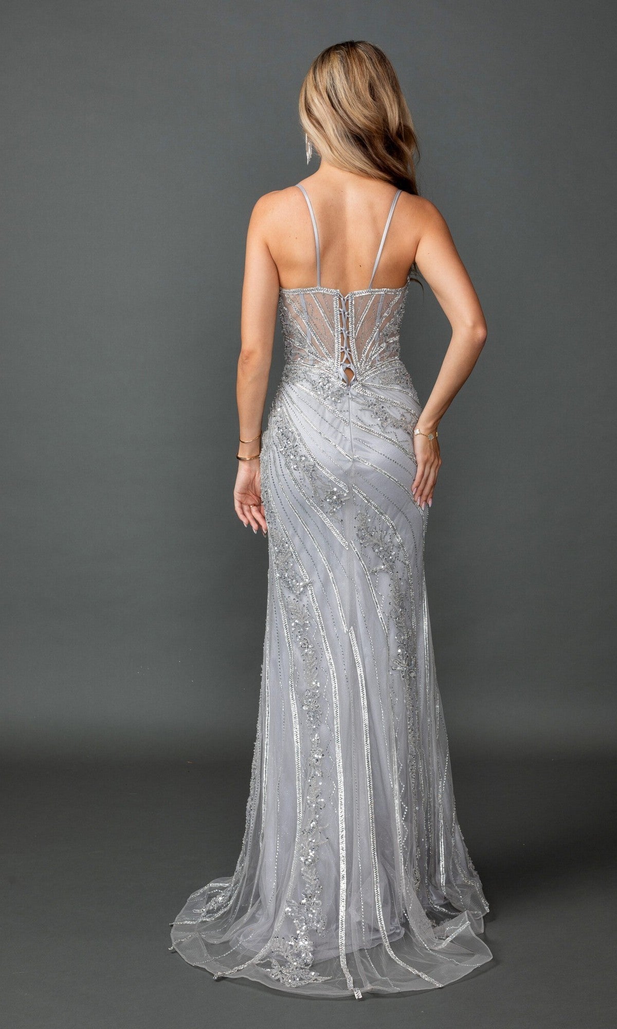 A woman is shown from the back, standing against a gray background, wearing the Dancing Queen 4599 long prom dress—a silver, floor-length sheath gown featuring thin straps and sparkling sequin detailing.