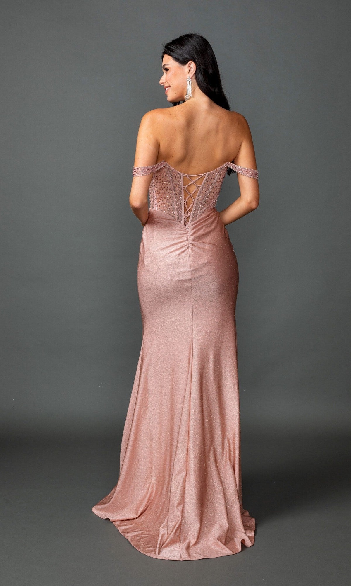 A woman stands against a plain gray background, facing away from the camera, wearing the Dancing Queen 4601 long prom dress in blush pink—an off-the-shoulder gown with a corset bodice, lace-up back, beaded details, and a high-leg slit.