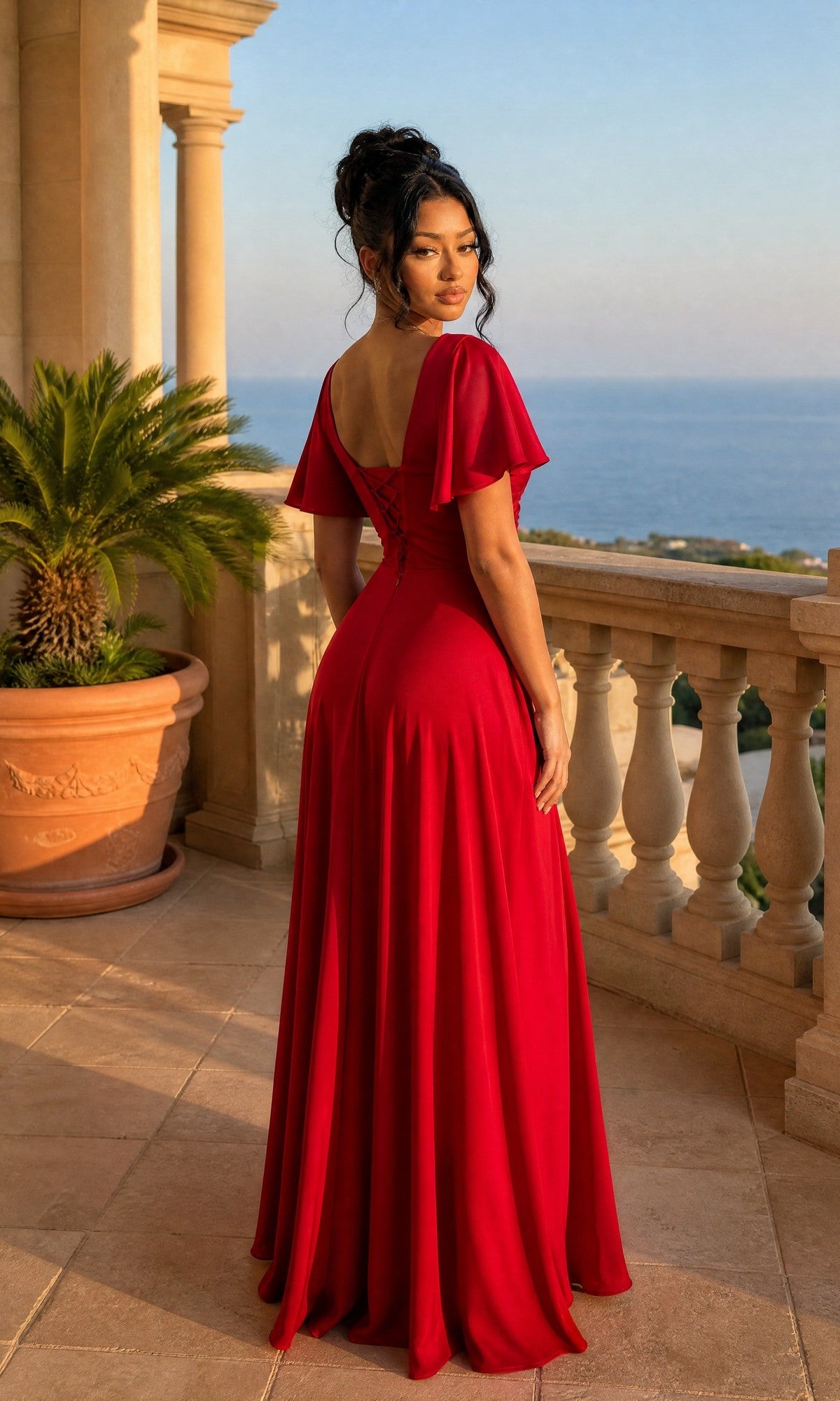 Wearing the Dancing Queen 4602 long formal dress, a woman with her back turned stands on a balcony overlooking the sea, dressed in red with flutter sleeves and a potted palm plant nearby.