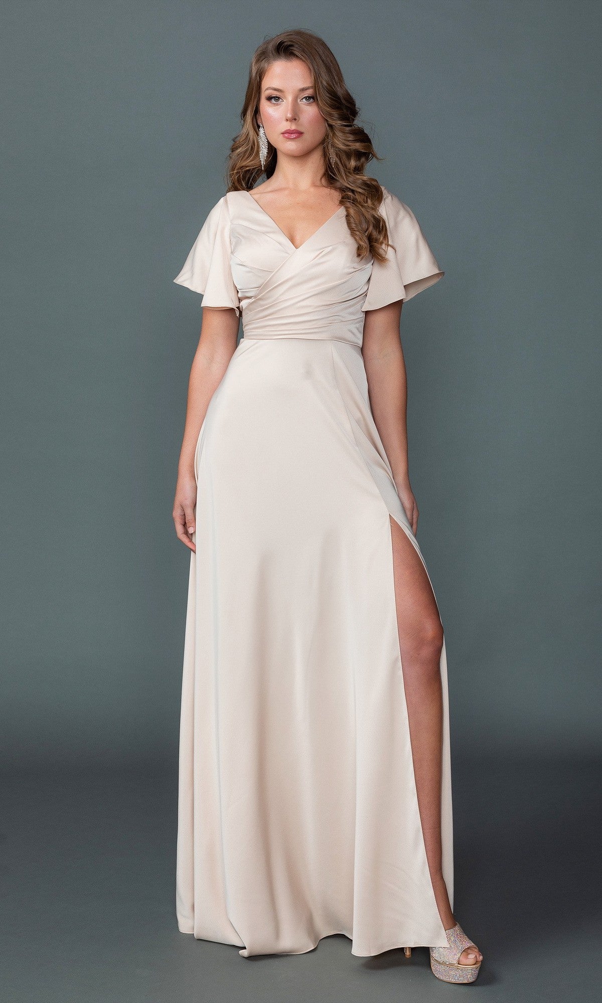 A woman in the Dancing Queen 4602 long formal dress in champagne, featuring a v-neckline, short flutter sleeves, wrapped bodice, and high side slit, stands against a plain gray background.