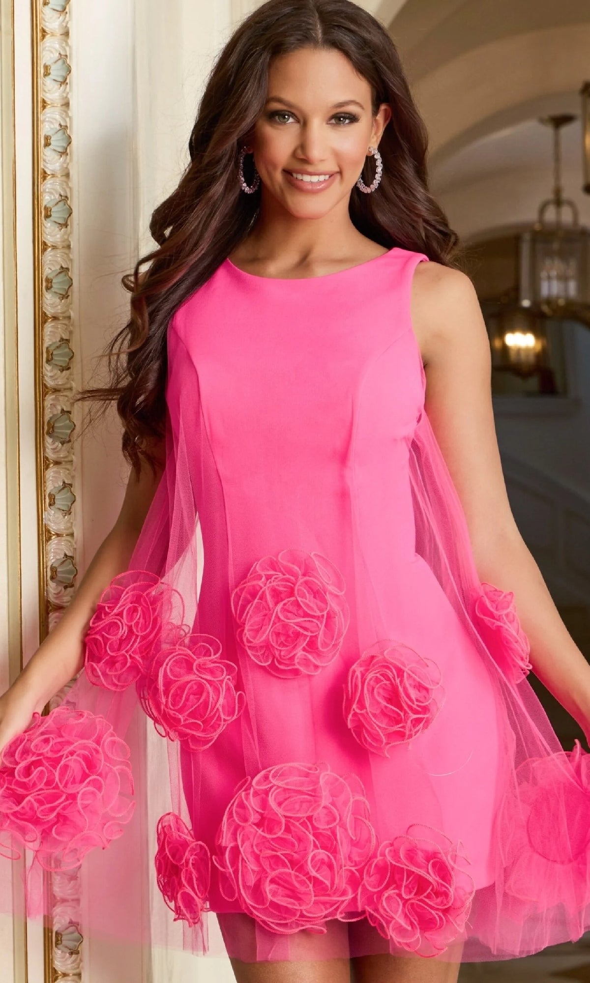 A woman in the Jovani 46135 Short Rosette Hoco Dress, a sleeveless bright pink dress with large fabric flower accents and mesh panels, stands indoors and smiles at the camera.
