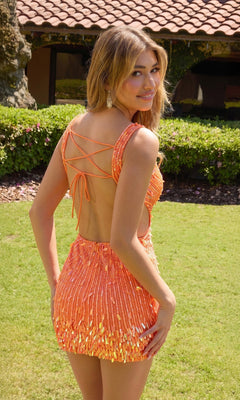 A woman stands on grass outdoors, looking over her shoulder, wearing the sparkly orange Primavera 4617 short homecoming dress with crisscross straps. Bushes and a tiled-roof building are in the background.