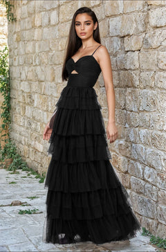 A woman stands outdoors by a stone wall, wearing the Blondie Nites 4637BN long black ruffled prom dress with thin straps, a sweetheart bodice, and a small cutout under the bust.