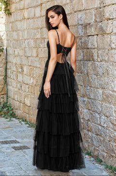 A woman in the Blondie Nites 4637BN Long Ruffled Prom Dress with a sleeveless, lace-up back stands on a stone walkway by a textured wall, glancing over her shoulder.