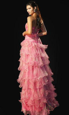 A woman stands against a black background wearing the Jovani 46408 long prom dress in watercolor pink, its strapless, floor-length ruffle organza gown cascading as she looks over her shoulder.
