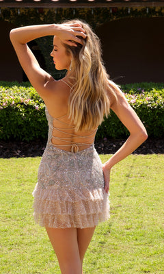 A woman stands on grass with her back to the camera, one hand touching her hair, wearing the sparkly Primavera 4647 Short Homecoming Dress featuring an open back with lace-up detail and a ruffled skirt. Shrubs and a building appear in the background.