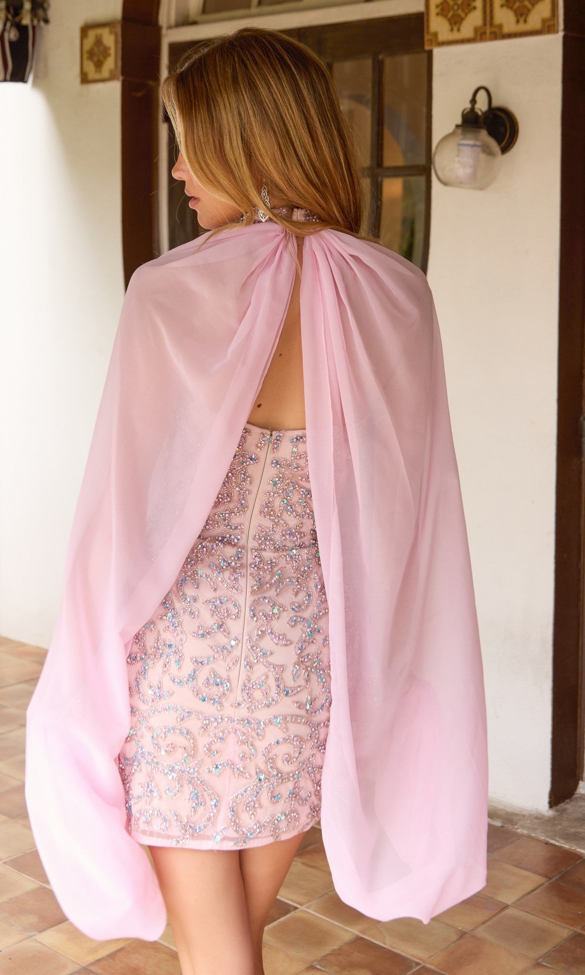 A woman stands indoors facing away from the camera, wearing the Primavera 4649 short homecoming dress—a light pink bodycon fitted design adorned with sequins and crystal embellishments, and featuring an attached sheer pink cape.