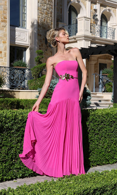 Wearing the Jovani 47000 long formal dress, a woman stands gracefully in a manicured garden before a grand stone building, her strapless gown featuring a gold floral waist detail as she holds the side of its pleated hot pink skirt.