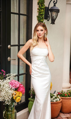 A woman in the Ava Presley 47026 long prom dress poses beside a door, surrounded by flower arrangements and potted plants.