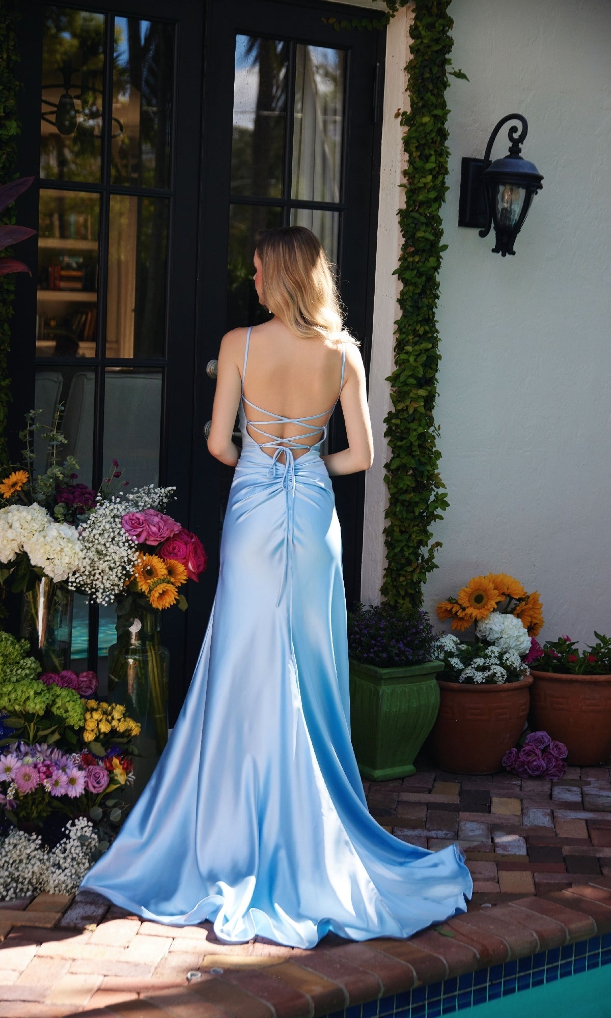 A woman in the Ava Presley 47030 Strappy-Back Long Prom Dress in light blue stands by a pool, facing away near glass doors, with flower arrangements and potted plants around her.