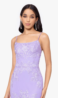 A woman wears the Blondie Nites Lilac Purple Long Prom Dress 4706BN, featuring thin straps and floral details, paired with long dangling earrings.