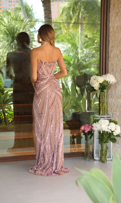 A woman in the Ava Presley 47123 Strapless Long Sequin Prom Gown stands facing a glass window, surrounded by potted and vase-arranged flowers in an outdoor garden.