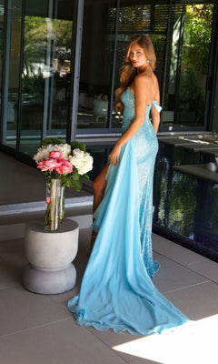 A woman stands indoors near a glass wall and a vase of flowers on a round table, wearing the Ava Presley 47124 long one-shoulder light blue prom dress.
