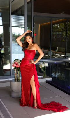 A woman in the Ava Presley 47124 long prom dress, shimmering red and sequined, holds a rose by the poolside patio with elegant floral arrangements on nearby tables.