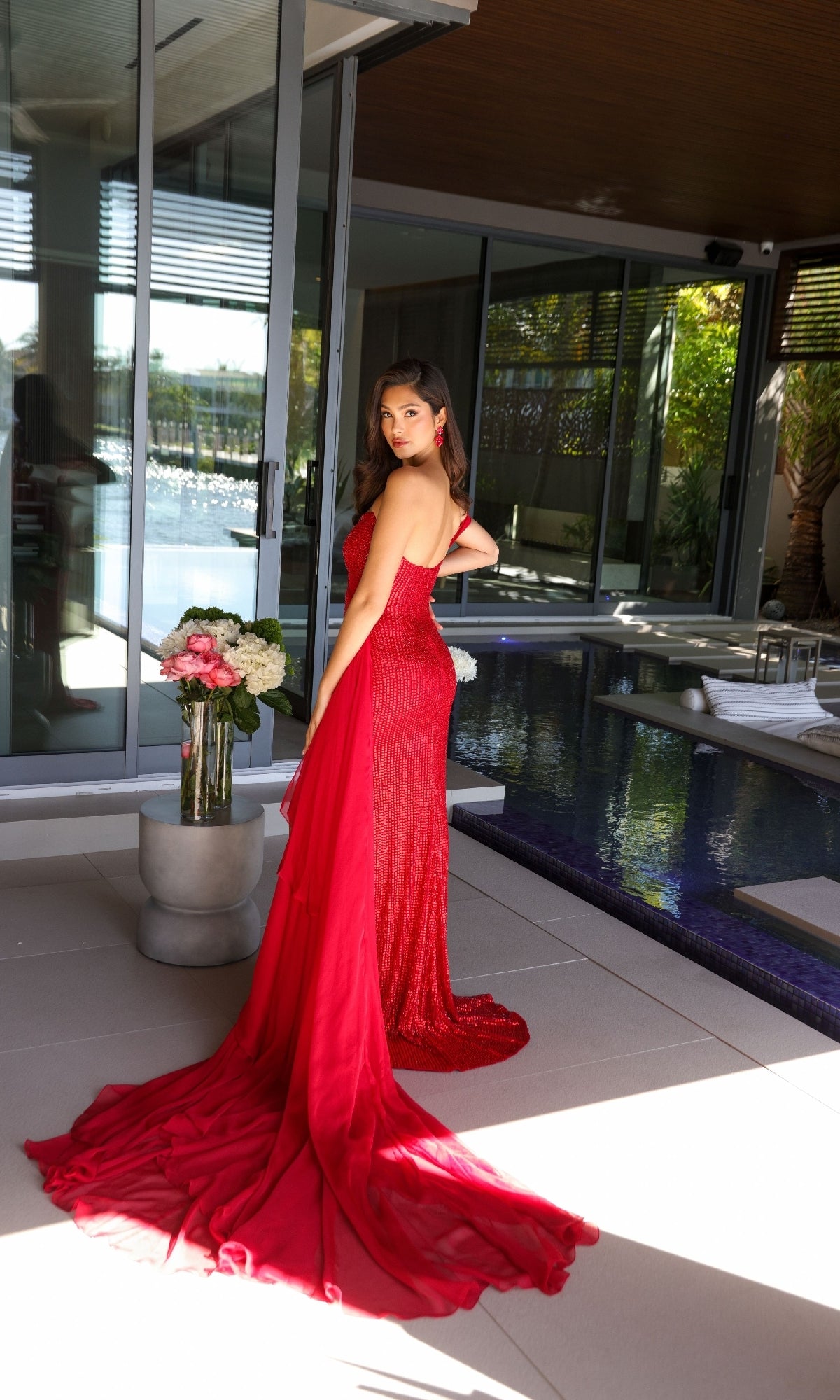 A woman in the Ava Presley 47124 long, red one-shoulder prom dress stands by an indoor pool, looking over her shoulder. A vase of flowers sits on a small table nearby.