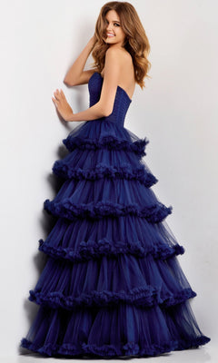 A woman stands against a light wall wearing the Jovani 47139, a long navy blue strapless prom dress with floor-length, tiered ruffles for an elegant look.