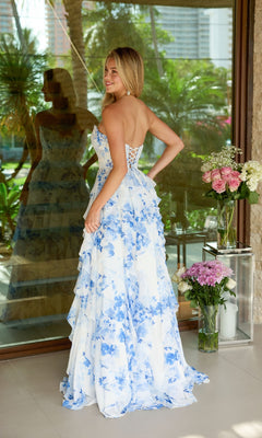 A woman models the Ava Presley 47145 long prom dress, featuring a strapless floral design and lace-up open back, as she stands indoors by vases of flowers with greenery visible through the window.