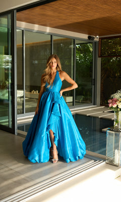 A woman in a vivid blue Halter Long Balloon Prom Dress, Ava Presley 47146, stands indoors near a modern pool, with flowers and glass doors behind her.