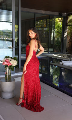 A woman in the Ava Presley 47147 long prom dress, featuring sequins and a thigh-high slit, stands near an indoor pool beside a bouquet of flowers, glancing over her shoulder.