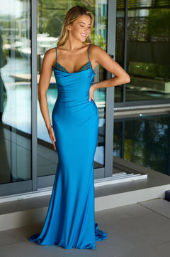 A woman stands by large windows wearing the Ava Presley 47173 long prom dress in turquoise, a fitted, floor-length stretch jersey gown with thin straps and beaded neckline embellishments.