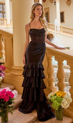 A woman wearing the Ava Presley 47176 long black prom dress with a tiered ruffle skirt and corset bodice stands by a stone railing, surrounded by flowers and with a fountain in the background.