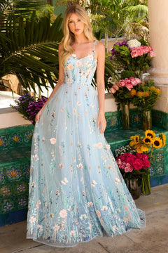 A woman stands outdoors in the Ava Presley 47189 long prom dress, featuring a light blue floral design with a corset bodice, thin straps, and a flowing A-line skirt amid colorful flowers and lush greenery.
