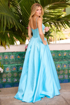 Wearing the Ava Presley 47192 long, light blue prom dress with thin straps and a corset bodice, a woman stands outdoors near green tiles and tropical plants, holding a bouquet and facing away.
