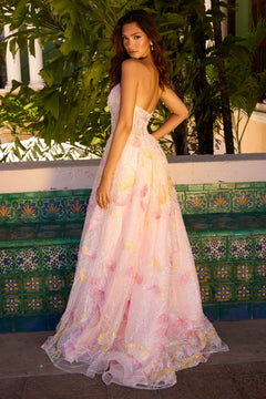 A woman stands outdoors in the Ava Presley 47193 Strapless Pink Floral Prom Gown, its floor-length design featuring yellow floral patterns, as she looks over her shoulder at the camera.