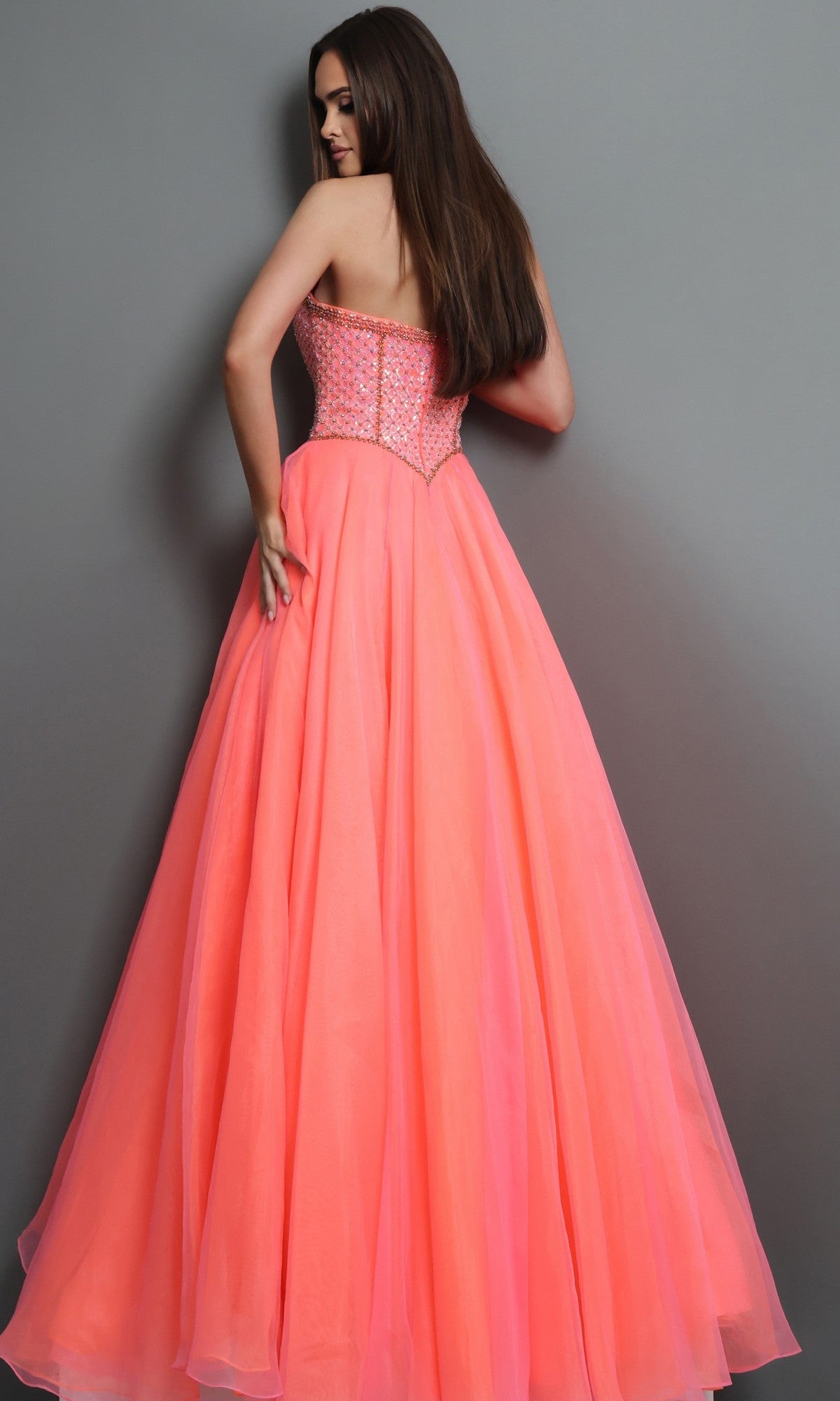 A woman stands with her back to the camera against a plain gray background, wearing the Jovani 47196 Long Formal Dress: a floor-length, strapless designer gown in coral pink, featuring a fitted, beaded bodice and a flowing skirt.