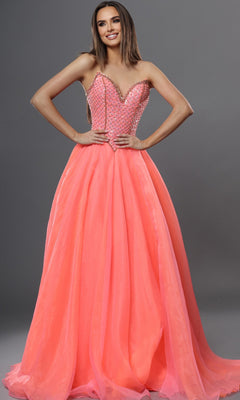 A woman stands in a studio wearing the Jovani 47196 long formal dress—a strapless, beaded ball gown in neon orange with a fitted bodice and a full, flowing skirt—set against a plain gray background.