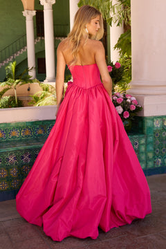 A woman wearing the Ava Presley 47204 long prom dress stands outdoors by a tiled wall and flower arrangement, facing away from the camera.