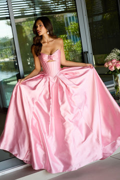 A woman indoors holds the sides of her pink Strapless Satin Prom Ball Gown (Ava Presley 47223), with a bouquet of pink roses on a table beside her.