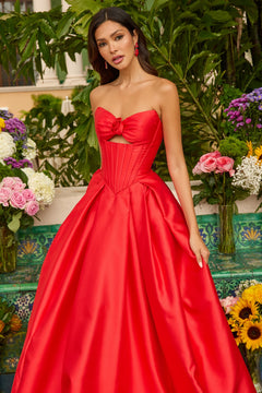 A woman stands before lush greenery and vibrant flowers, wearing the Ava Presley 47223 strapless satin prom ball gown in bright red with a bow detail on the bodice.