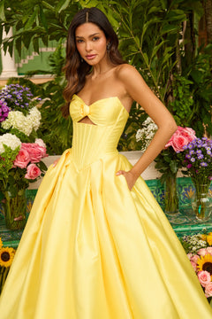 A woman wears the Ava Presley 47223 Strapless Satin Prom Ball Gown, featuring a bow detail, as she poses in a garden surrounded by colorful flowers and lush greenery.