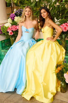 Two women in Ava Presley 47223 strapless satin prom ball gowns—one light blue, one yellow—sit side by side, surrounded by colorful flowers and lush greenery.