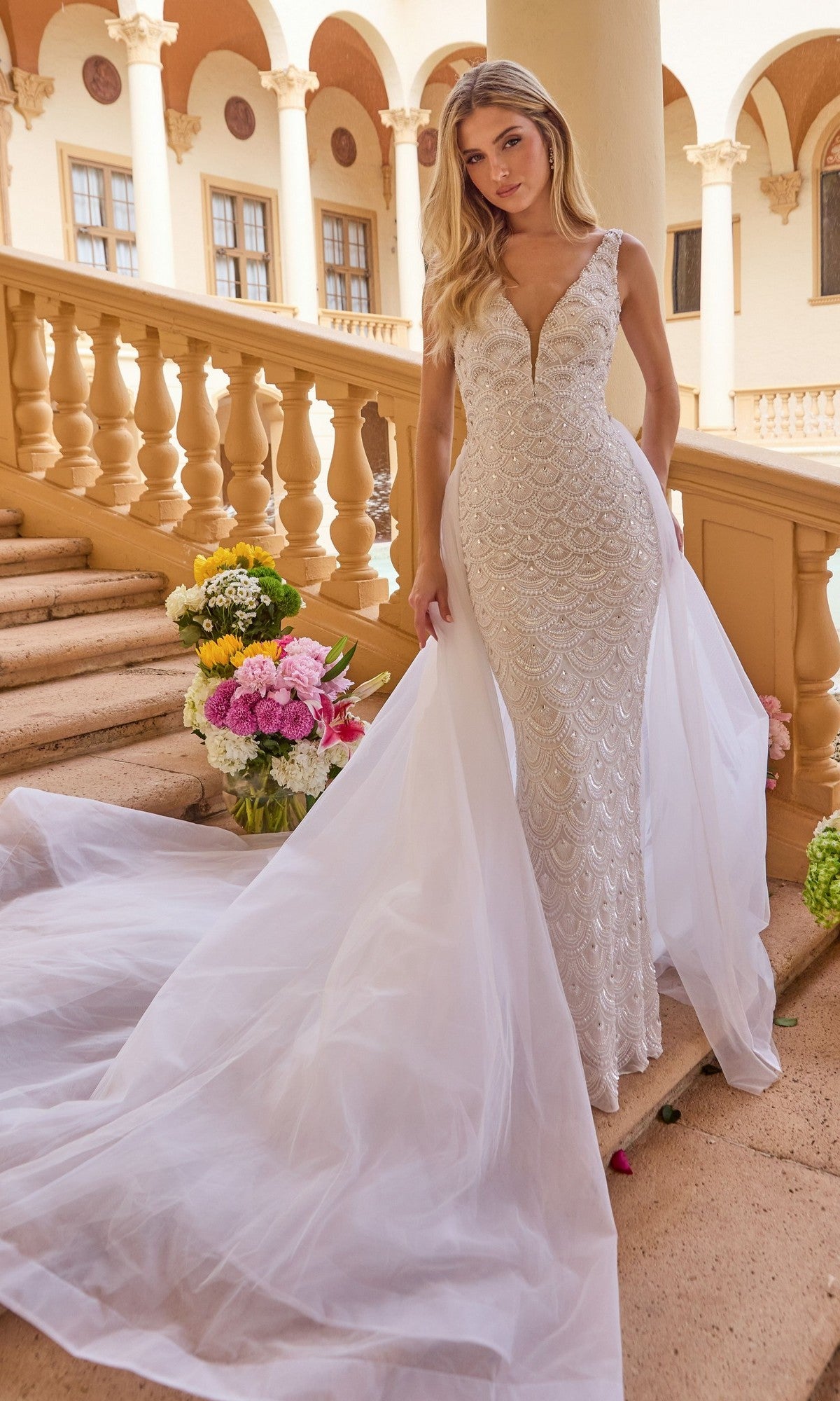 A woman stands on stone steps in the Ava Presley 47224, a fitted, beaded white long formal dress with an open back and tulle overskirt. Flower arrangements and ornate columns and arches frame the elegant scene.