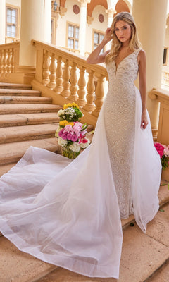 A woman in the Ava Presley 47224 long formal dress—a sleeveless, beaded white gown with an open back and extended tulle train—stands on stone stairs next to vibrant flower arrangements.