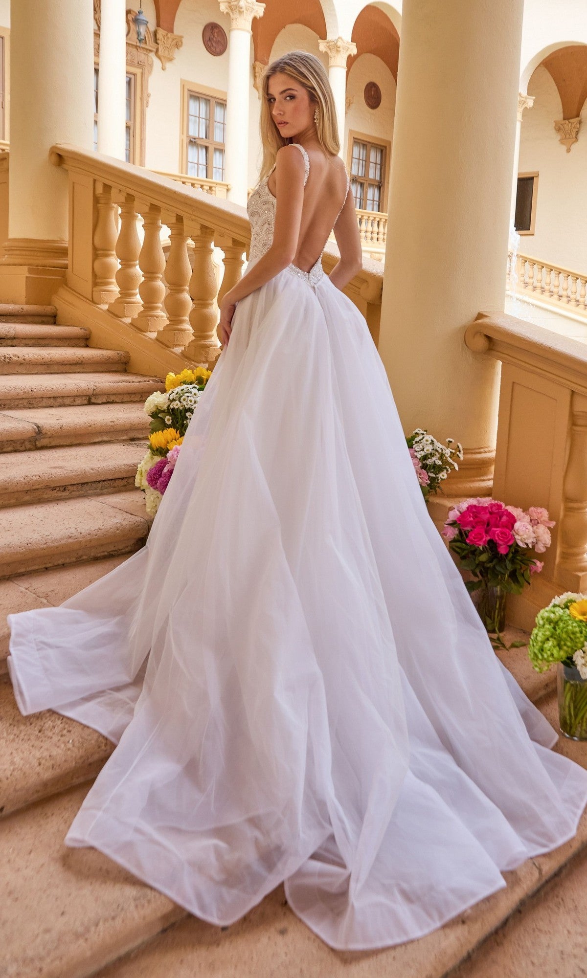 A woman wearing the Ava Presley 47224 long formal dress with a flowing tulle overskirt stands on stone stairs among floral arrangements in an outdoor architectural setting with columns and arches.
